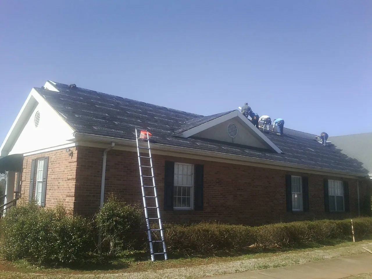 Crew tearing shingles off a brick ranch roof with a ladder set — Metal Roof Repair work by our crew in Keswick
